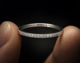 May include: A silver eternity ring, featuring a continuous line of small, clear gemstones. The ring is held between two fingers against a dark backdrop, highlighting the sparkling stones and the ring's delicate design. The ring is a piece of jewellery.