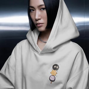 Op de afbeelding: Een witte hoodie met een kleurrijk cartoonontwerp op de voorkant. Het ontwerp bevat een zwart, pluizig personage, een gele ster en een ronde, paars-handvat lolly met een gezicht. Het model draagt een zilveren ketting.