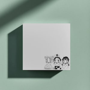 Könnte beinhalten: Ein weißer quadratischer Notizblock mit einer Schwarz-Weiß-Illustration von drei Anime-Charakteren. Die Charaktere haben Schleifen im Haar und tragen gemusterte Kleidung. Der Notizblock liegt auf einer hellgrünen Oberfläche.