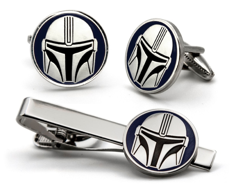 May include: A silver tie clip and cufflinks set featuring a Mandalorian helmet design. The helmet is silver with black details and a blue background.