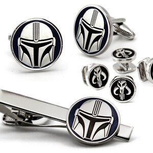 May include: A silver tie clip and cufflinks set featuring a Mandalorian helmet design. The tie clip has a silver bar with a black and silver Mandalorian helmet design. The cufflinks are round with a black and silver Mandalorian helmet design. The cufflinks have a silver back.