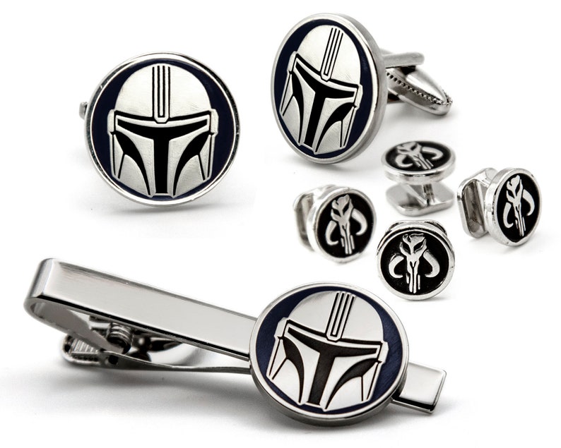 May include: A silver tie clip and cufflinks set featuring a Mandalorian helmet design. The tie clip has a silver bar with a black and silver Mandalorian helmet design. The cufflinks are round with a black and silver Mandalorian helmet design. The cufflinks have a silver back.
