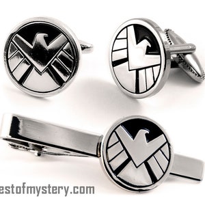 Può includere: Un set di gemelli e fermacravatta argentati. Ogni pezzo presenta un emblema circolare con un design nero e argento di un uccello stilizzato e forme geometriche. Il sito web chestofmystery.com è stampato in basso.