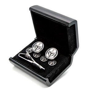 May include: A black gift box with a black velvet interior, containing a silver tie clip and cufflinks with a black and white Mandalorian helmet design.