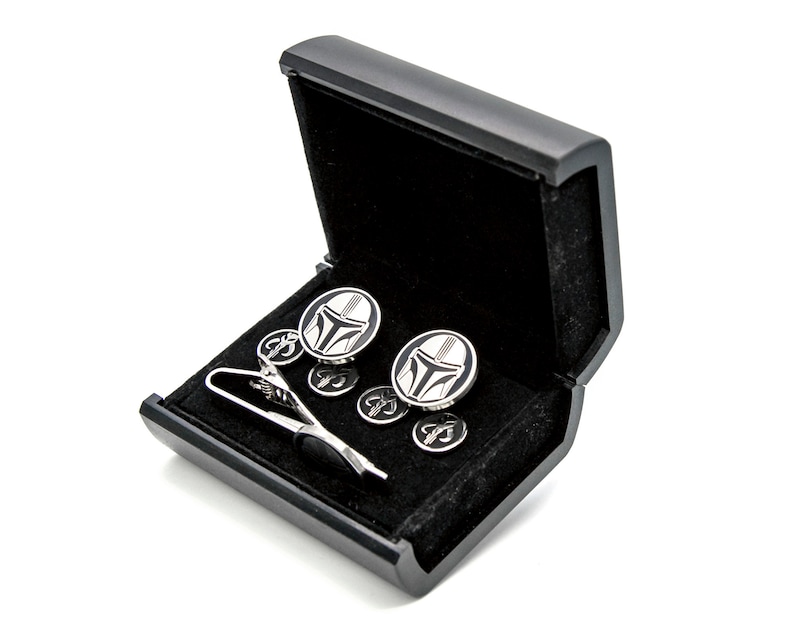 May include: A black gift box with a black velvet interior, containing a silver tie clip and cufflinks with a black and white Mandalorian helmet design.