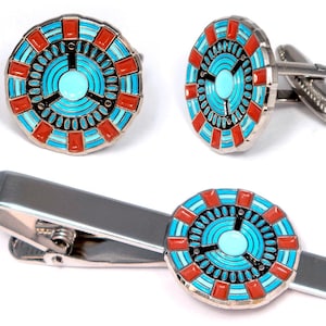 Può includere: Un set di gemelli e un fermacravatta in metallo color argento. Ogni pezzo presenta un design circolare con un cerchio azzurro al centro, circondato da anelli concentrici neri e azzurri e un bordo esterno con accenti rettangolari rossi.