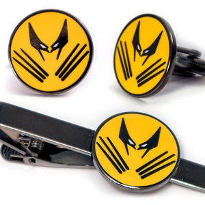 Puede incluir: Un conjunto de dos gemelos redondos y un pasador de corbata en metal gris oscuro. Cada pieza presenta una incrustación de esmalte amarillo con un diseño gráfico negro que representa una máscara estilizada y tres marcas de garras.
