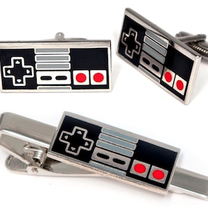 May include: A set of two rectangular cufflinks and one tie clip, each featuring a black, grey, and red enamel design modeled after a vintage video game controller with a directional pad, two grey buttons, and two circular red action buttons.