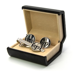 May include: A black gift box with a beige interior, containing a silver tie clip and cufflinks with a black and silver Mandalorian helmet design.