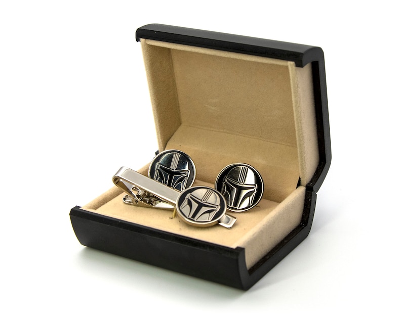 May include: A black gift box with a beige interior, containing a silver tie clip and cufflinks with a black and silver Mandalorian helmet design.