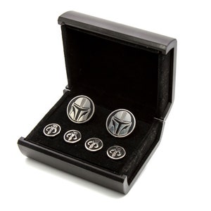 May include: A black wooden box with a velvet interior, containing a set of cufflinks and shirt studs. The cufflinks feature a silver Mandalorian helmet design, while the shirt studs feature a silver Mandalorian symbol.