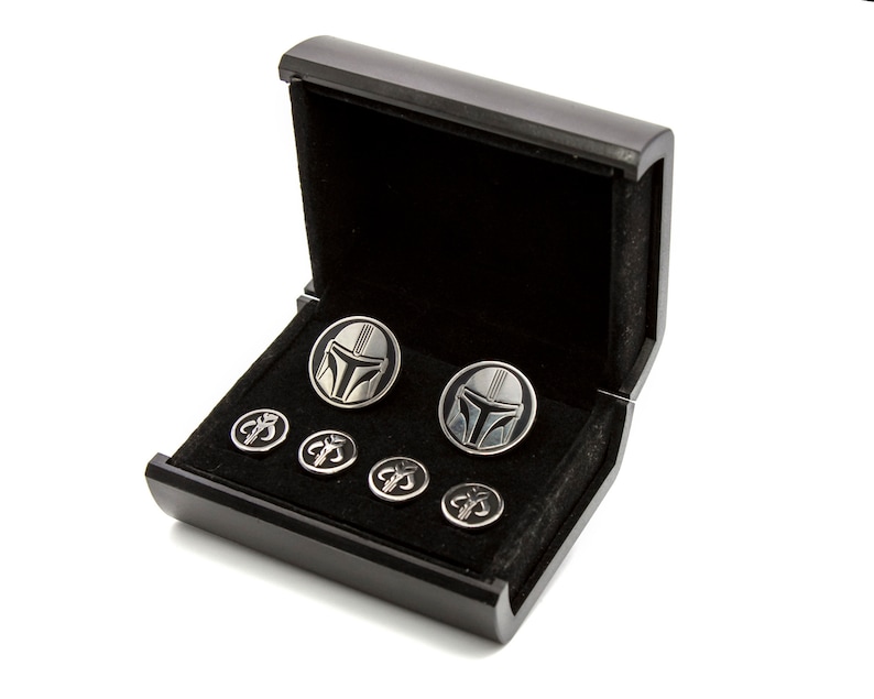 May include: A black wooden box with a velvet interior, containing a set of cufflinks and shirt studs. The cufflinks feature a silver Mandalorian helmet design, while the shirt studs feature a silver Mandalorian symbol.