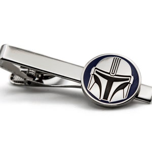 May include: A silver tie clip with a circular design featuring a black and white Mandalorian helmet on a blue background.