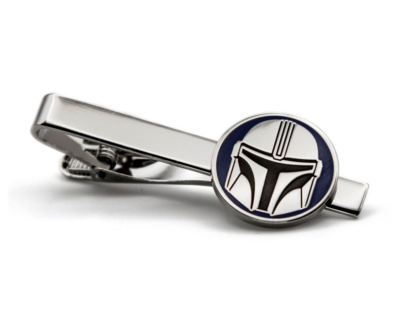 May include: A silver tie clip with a circular design featuring a black and white Mandalorian helmet on a blue background.