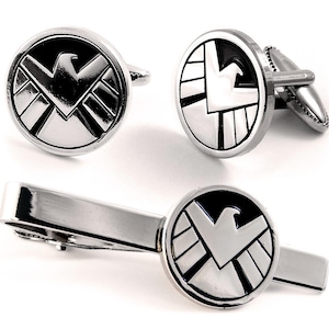 May include: A set of two circular cufflinks and one matching tie bar, all featuring a polished silver-tone finish with a black enamel inlay. Each piece displays a stylized eagle emblem centered within a circular border, creating a cohesive set.