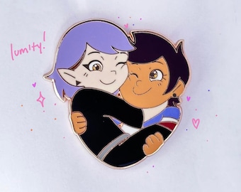 May include: A heart-shaped pin featuring two cartoon characters embracing. One character has purple hair and the other has brown hair. The pin has a gold border and the word "lumity!" is written in pink.