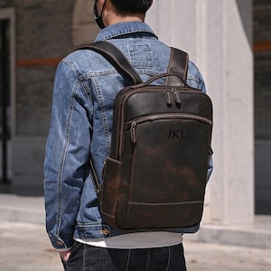 Op de afbeelding: Een bruine leren rugzak met een voorvak met rits en een groot compartiment. De rugzak heeft een bruine leren band en een zwart label met de tekst "JKL" aan de voorkant.