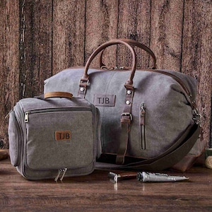 May include: A gray canvas travel duffel bag with brown leather accents and a personalized name tag that reads "TJB". The bag has a zipper closure and a shoulder strap. A smaller gray canvas toiletry bag with brown leather accents and a personalized name tag that reads "TJB" is also included.