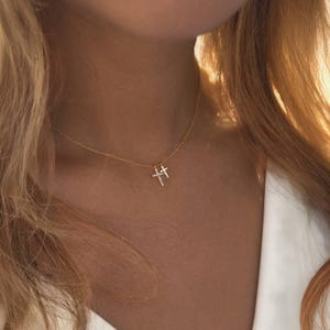 May include: A delicate gold necklace featuring two cross pendants. One cross is adorned with small, sparkling crystals. The necklace is worn against a background of blonde hair and a white garment.