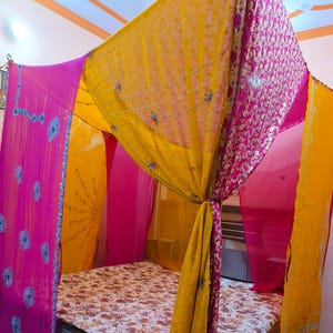 Może przedstawiać: Łóżko z baldachimem wykonanym z przezroczystych, kolorowych tkanin. Baldachim ma żywe różowe, żółte i złote zasłony, ozdobione haftowanymi detalami. Łóżko ma narzutę w kwiatowy wzór i jasną drewnianą ramę.