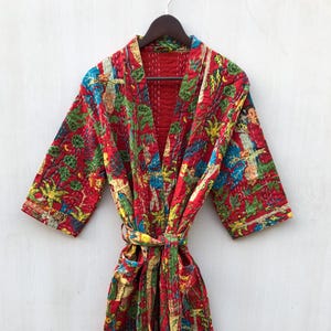May include: A red, quilted robe with a colorful, patterned design featuring figures and foliage. The robe has a matching belt and is hanging on a wooden hanger. The interior lining is a solid red color.