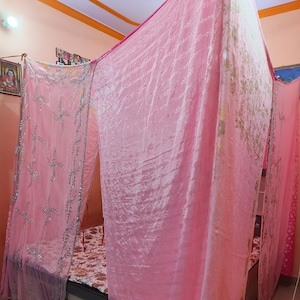 May include: A pink canopy bed with draped fabric. The bed frame is visible beneath the sheer, flowing curtains. The curtains are a soft pink colour, with silver embroidery on the side panels. The bedspread has a floral pattern.