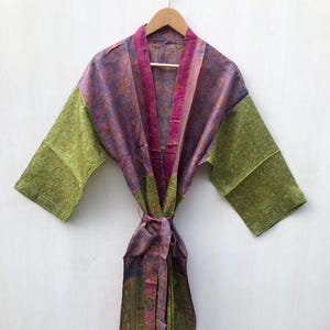Op de afbeelding: Een kleurrijke, patroonrijke kimono met een striksluiting. De kimono heeft een mix van paarse, roze en groene tinten. De mouwen zijn groen en de kraag en strik zijn roze. De kimono hangt aan een houten hanger.