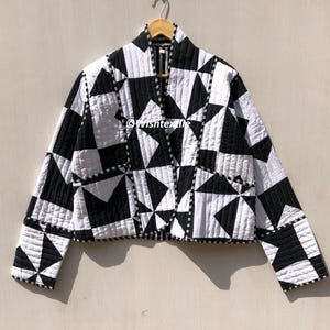 May include: Black and white quilted jacket with a geometric pattern. The jacket has a V-neck and long sleeves.  The jacket is cropped and has a relaxed fit.