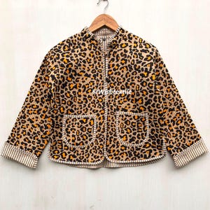 May include: A leopard print quilted jacket in tan, black, and orange. It features a stand-up collar, a front zip, and two front pockets. The cuffs and edges are trimmed with a striped fabric. The jacket is hung on a wooden hanger.