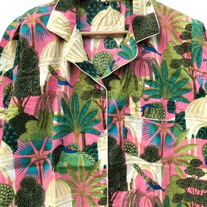 Pu&ograve; includere: Una camicia rosa con bottoni e bordino bianco. La camicia presenta una stampa colorata di pavoni, alberi ed edifici. La camicia ha una tasca sul lato sinistro.