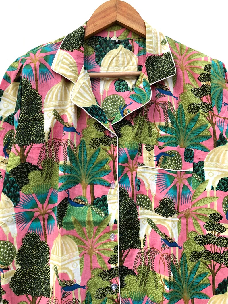 Pu&ograve; includere: Una camicia rosa con bottoni e bordino bianco. La camicia presenta una stampa colorata di pavoni, alberi ed edifici. La camicia ha una tasca sul lato sinistro.