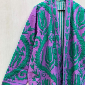May include: A purple embroidered jacket with a green floral design. The jacket is hanging on a wooden hanger. The interior lining is striped with green and white. The sleeves and front panels are covered in green floral embroidery.