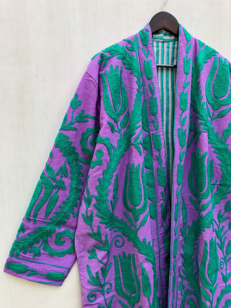 May include: A purple embroidered jacket with a green floral design. The jacket is hanging on a wooden hanger. The interior lining is striped with green and white. The sleeves and front panels are covered in green floral embroidery.