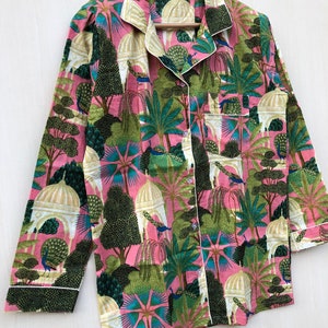 Pu&ograve; includere: Una camicia rosa a maniche lunghe con un bordo bianco e una stampa colorata di pavoni, alberi ed edifici.
