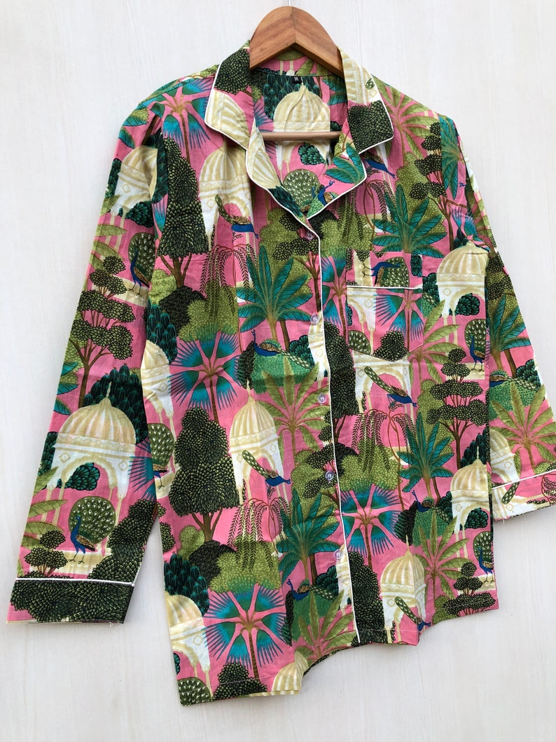 Pu&ograve; includere: Una camicia rosa a maniche lunghe con un bordo bianco e una stampa colorata di pavoni, alberi ed edifici.