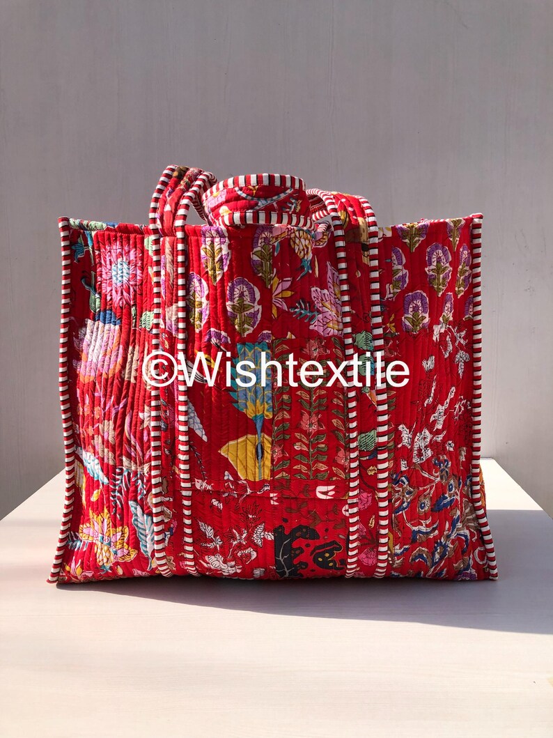 May include: A quilted tote bag in a vibrant red fabric featuring a multi-coloured floral pattern. The bag has two sturdy handles and edges trimmed with a white and red striped fabric. The text Wishtextile is printed across the centre.