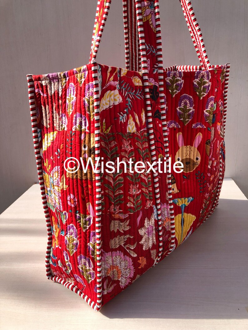 May include: A quilted tote bag in a vibrant red fabric featuring a repeating pattern of stylised flowers, leaves, and a small rabbit. The bag has red and white striped fabric trim along the edges and handles. The text Wishtextile is visible.