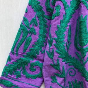May include: Close-up of a purple garment with intricate green embroidered floral and leaf patterns. The embroidery is detailed and covers the fabric, creating a textured design. The sleeves are visible, showcasing the repeating design.