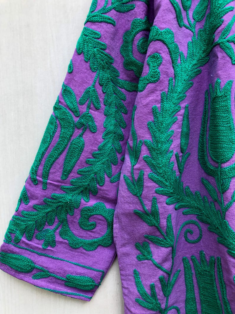May include: Close-up of a purple garment with intricate green embroidered floral and leaf patterns. The embroidery is detailed and covers the fabric, creating a textured design. The sleeves are visible, showcasing the repeating design.