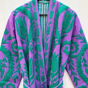 May include: A purple robe with green floral embroidery. The robe has a matching belt and a striped lining. The robe is hanging on a wooden hanger.
