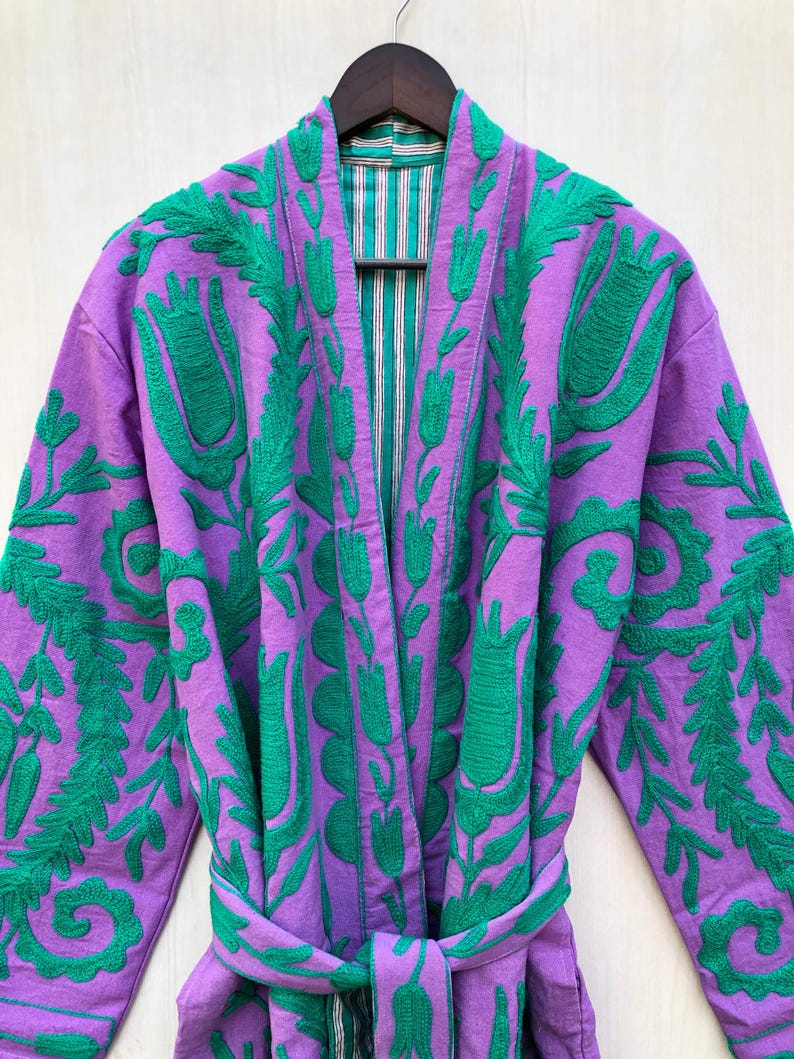 May include: A purple robe with green floral embroidery. The robe has a matching belt and a striped lining. The robe is hanging on a wooden hanger.