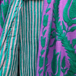 May include: A purple jacket with green floral embroidery and a striped teal and white lining. The jacket is hanging on a black hanger, showcasing the contrasting colors and textures of the garment.