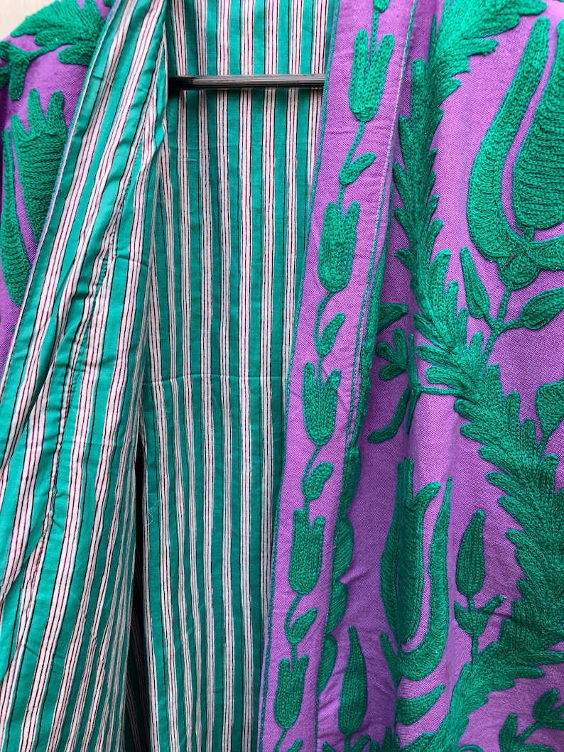 May include: A purple jacket with green floral embroidery and a striped teal and white lining. The jacket is hanging on a black hanger, showcasing the contrasting colors and textures of the garment.