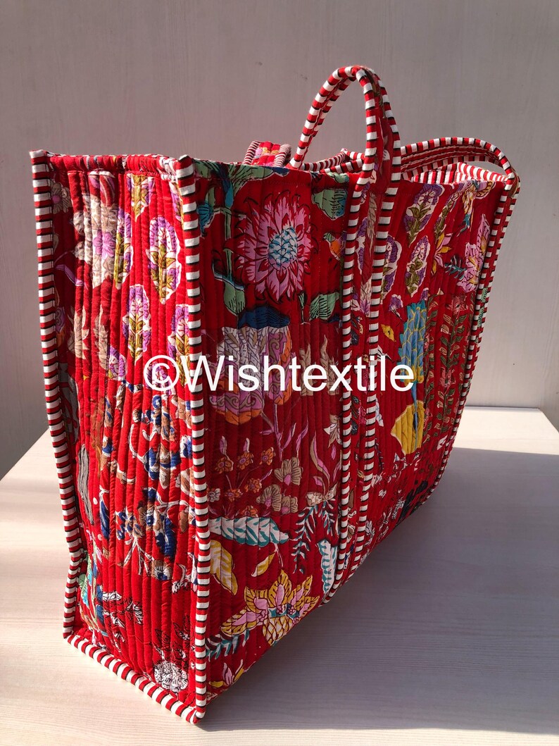 May include: A quilted tote bag featuring a vibrant red base with a multi-coloured floral pattern. The bag has white and red striped fabric trim along the edges and handles. The text "Wishtextile" is printed in white across the centre of the bag.