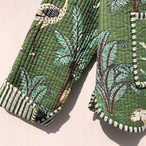 May include: Green quilted jacket with a white and green tropical print featuring palm trees, birds, and a leopard. The jacket has white and green striped trim.