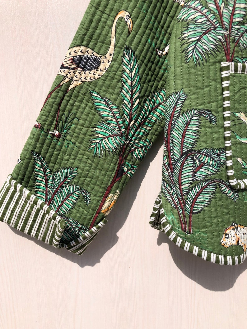 May include: Green quilted jacket with a white and green tropical print featuring palm trees, birds, and a leopard. The jacket has white and green striped trim.