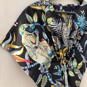 Puede incluir: Kaftán negro con un estampado botánico y de monos vibrante. El diseño presenta acentos amarillos, azules y blancos sobre un fondo oscuro. El kaftán está colgado de una percha de madera.