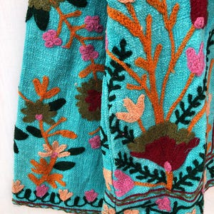 May include: A turquoise fabric with colorful embroidered flowers and leaves. The fabric has a textured surface and a fringed edge.