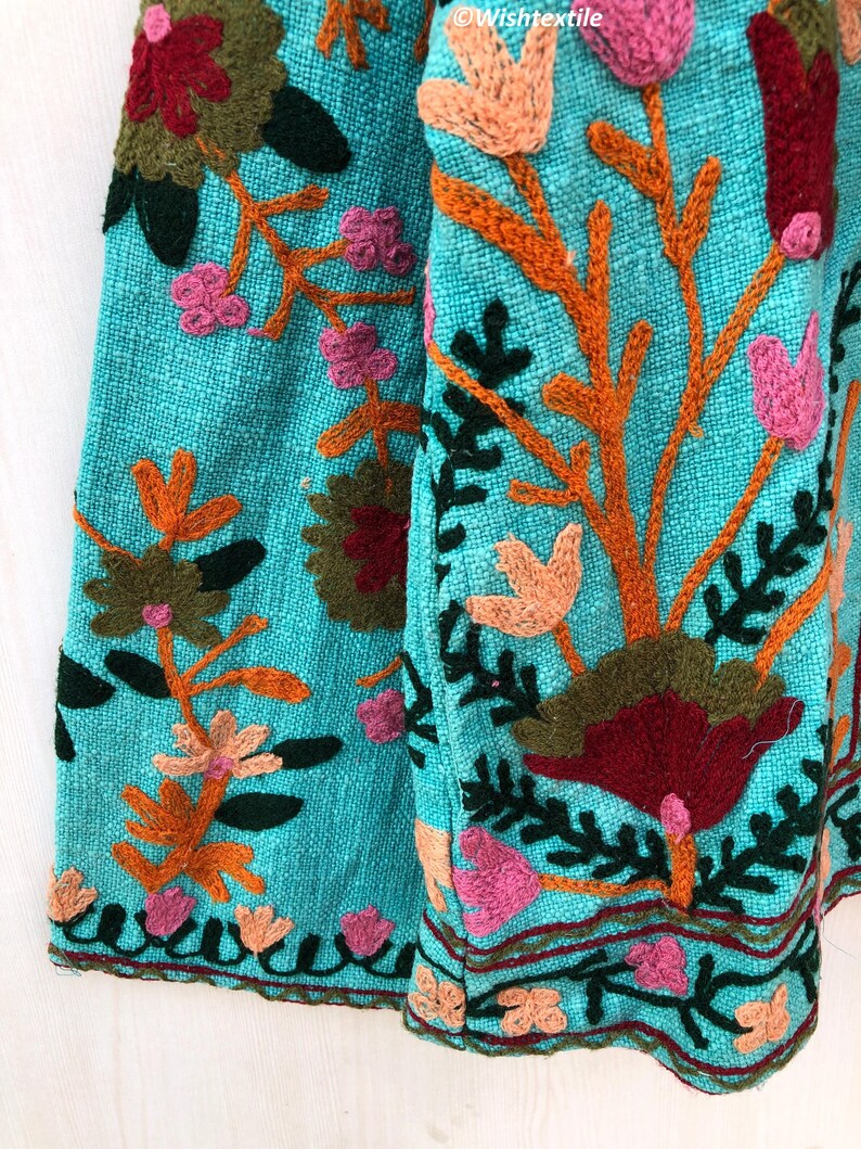 May include: A turquoise fabric with colorful embroidered flowers and leaves. The fabric has a textured surface and a fringed edge.