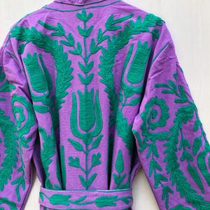 May include: A purple robe with green floral embroidery. The robe features a shawl collar, long sleeves, and a matching belt. The embroidery is detailed and covers the back and sleeves of the robe.